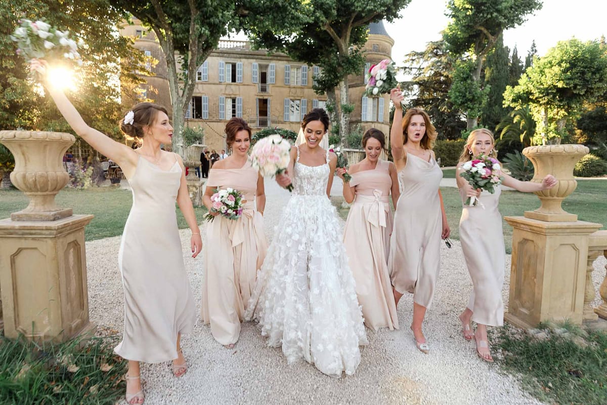 Bride and five bridesmaids in champagne satin dresses walking on gravel path at French chateau
