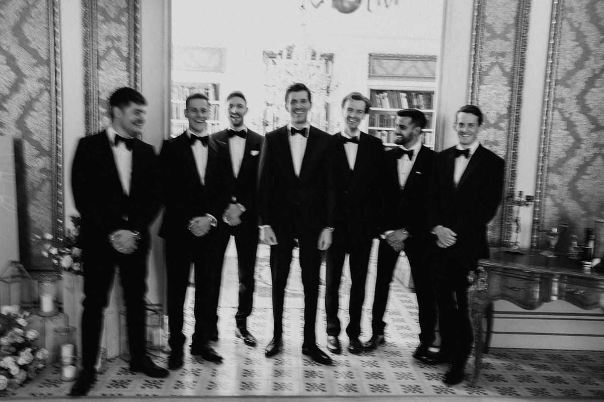 Black and white groom and six groomsmen in tuxedos lined up in chateau interior with patterned panels and mirror