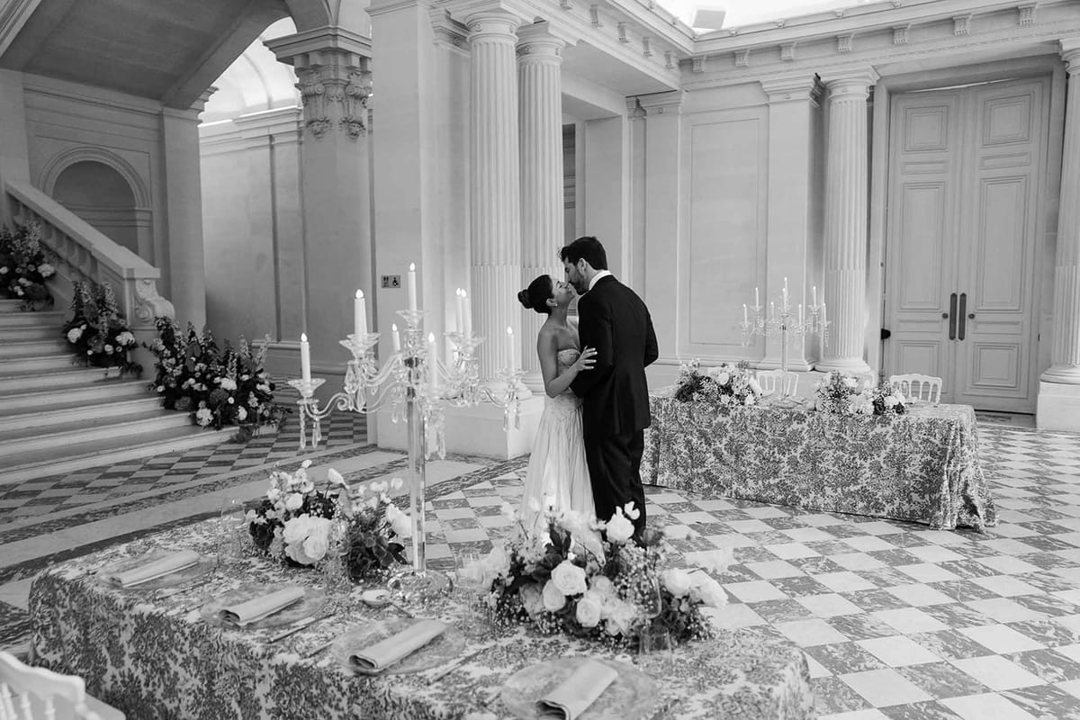 Black and white wide shot of bride and groom kissing between reception tables in a grand columned hall