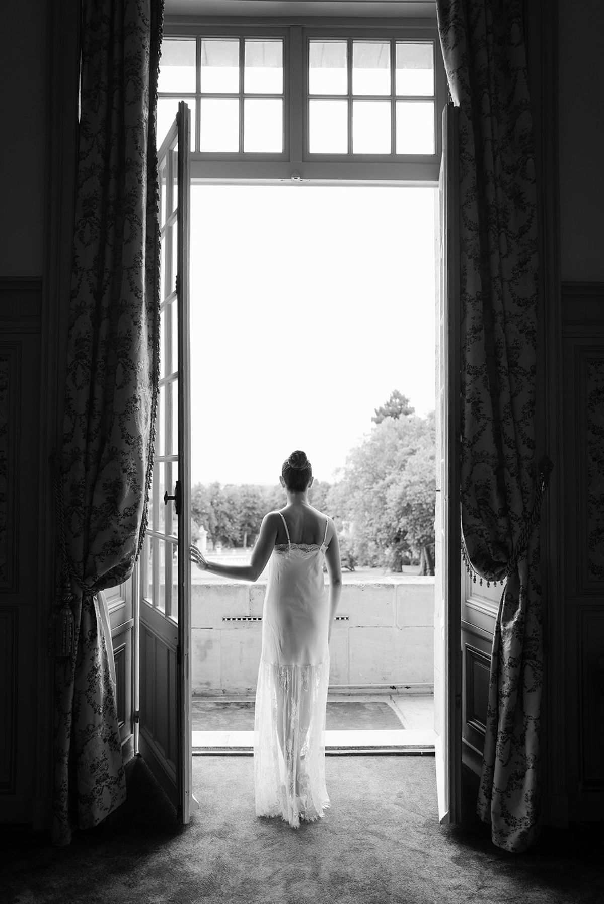 Black and white bride in spaghetti-strap lace gown standing at open French doors in chateau