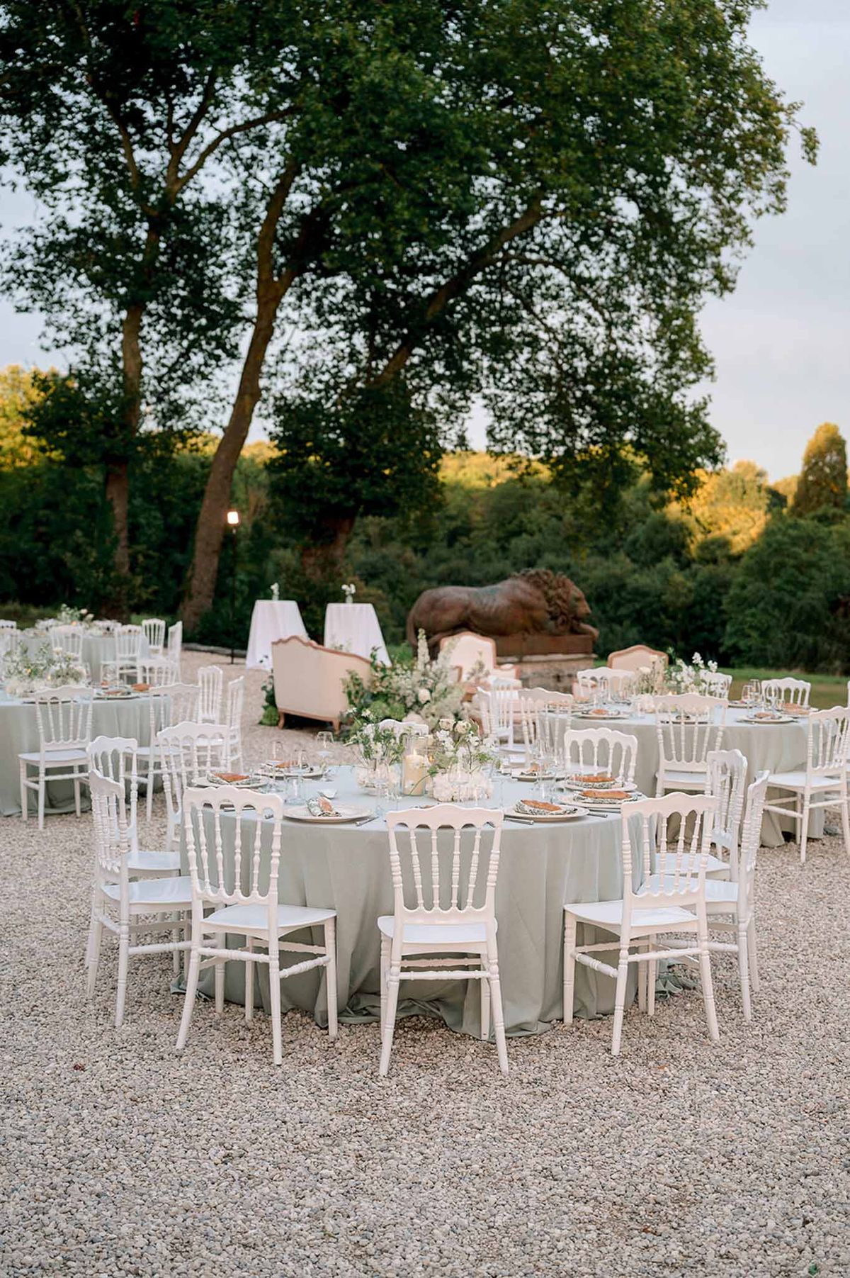 Outdoor reception with sage green linens, round tables, and bronze lion sculpture at dusk