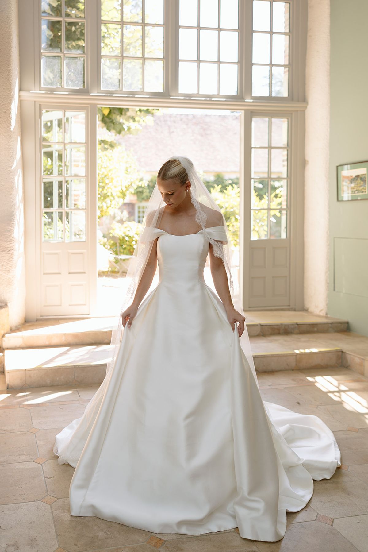 Bride in off-shoulder satin ball gown with lace-trimmed veil standing before open French doors