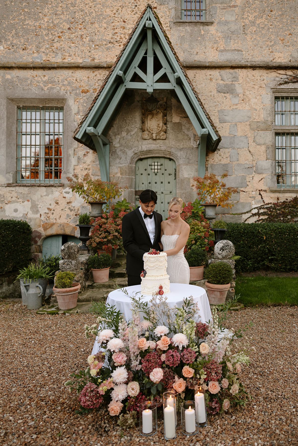 Bride and groom cutting fig-topped ivory cake surrounded by blush roses and candles before chateau