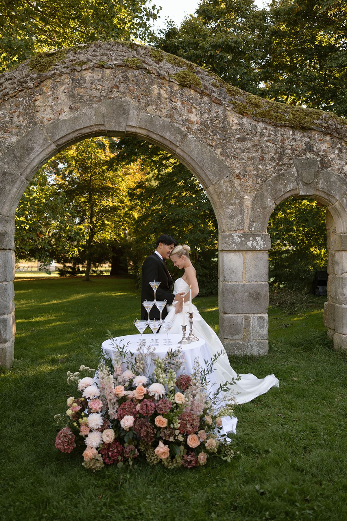 Bride and groom standing beneath a medieval stone arch behind a champagne tower table with peach and blush floral arrangement