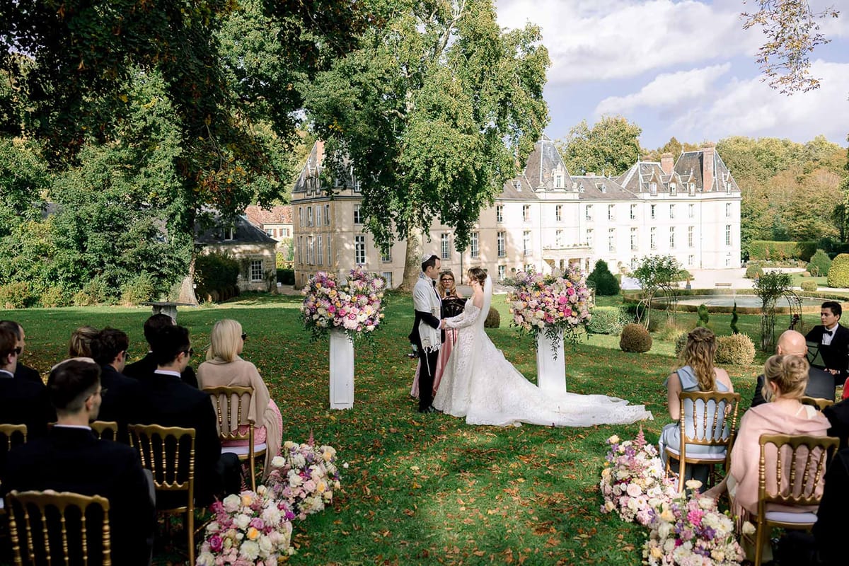 Jewish ceremony with pink and coral floral pedestals, gold Chiavari chairs, and chateau gardens behind