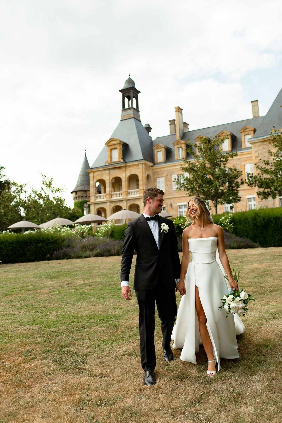 Laughing bride and groom walk hand in hand across chateau lawn with turrets in background