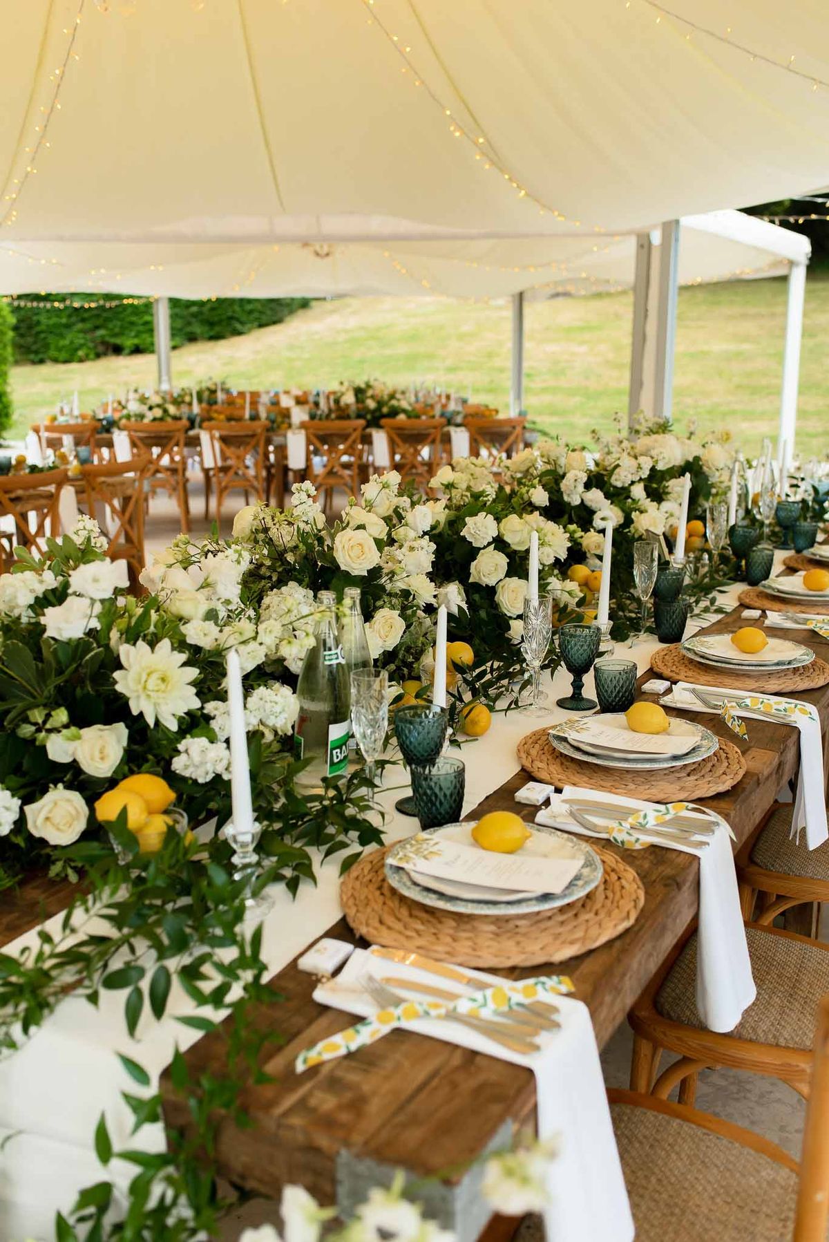 Lemon-themed farm table with citrus centerpiece and teal glassware under cream sailcloth tent with fairy lights