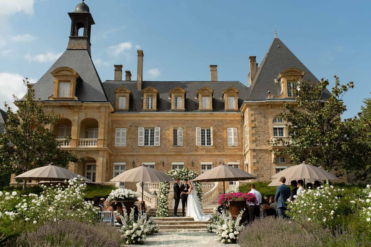 Couple at white floral arch before grand chateau staircase with lavender aisle and market umbrellas