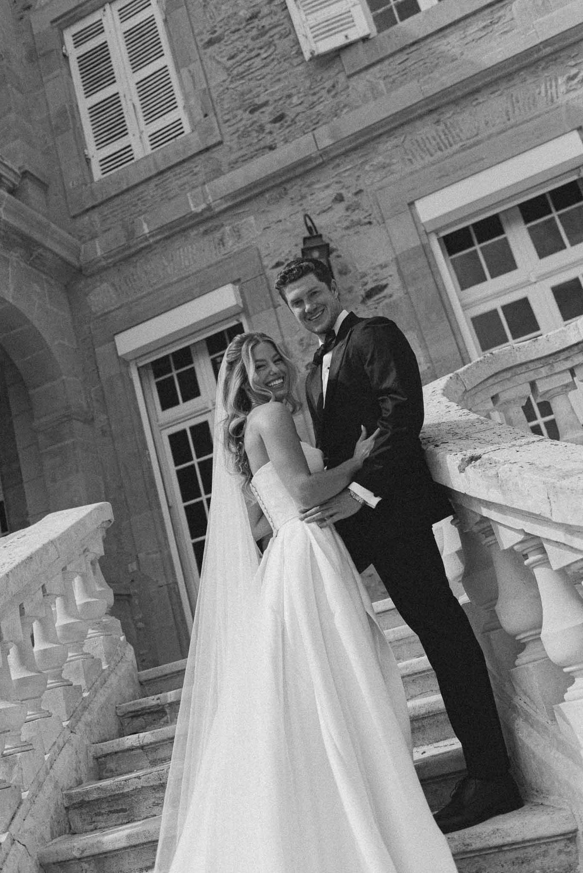 Laughing couple embraces on stone staircase with carved balustrade before chateau facade in B&W