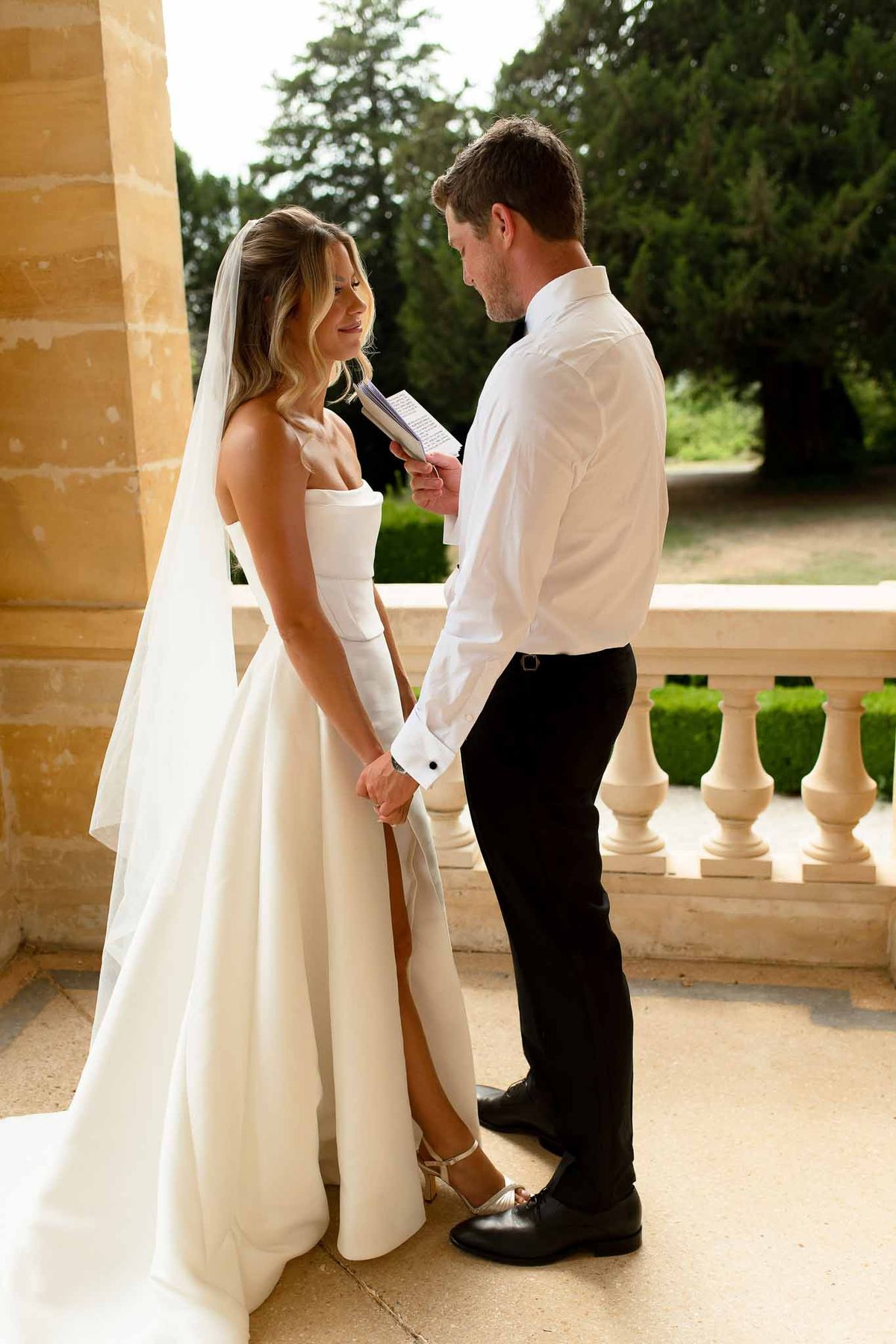 Groom reading vows from booklet holding bride's hand on stone balustrade terrace with conifers and hedgerows behind