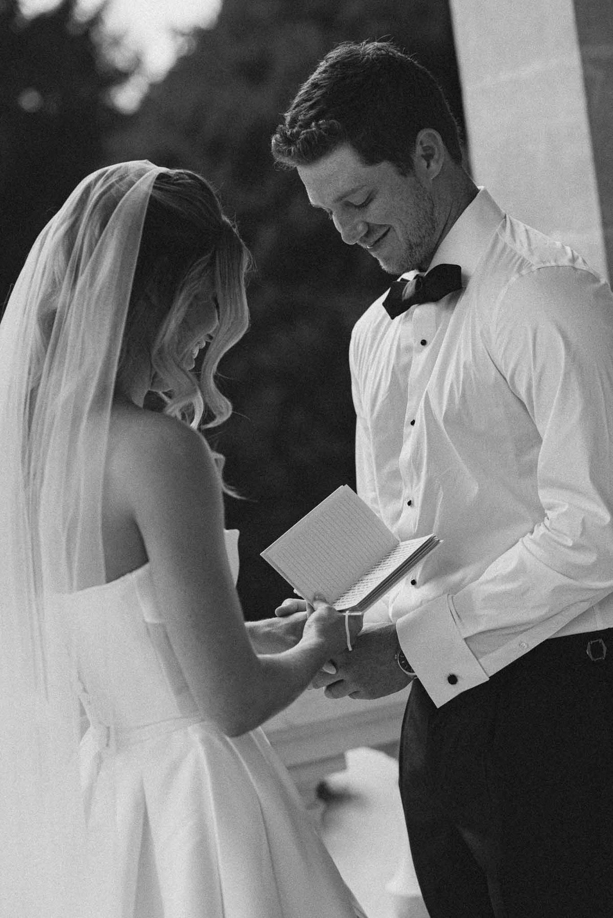 Black and white couple exchanging vows, groom holding notebook, bride in flowing veil with hands joined