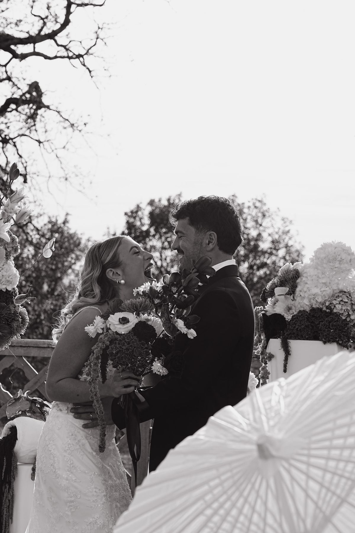 Bride and groom laugh nose to nose at the ceremony arch in black and white, bouquet between them