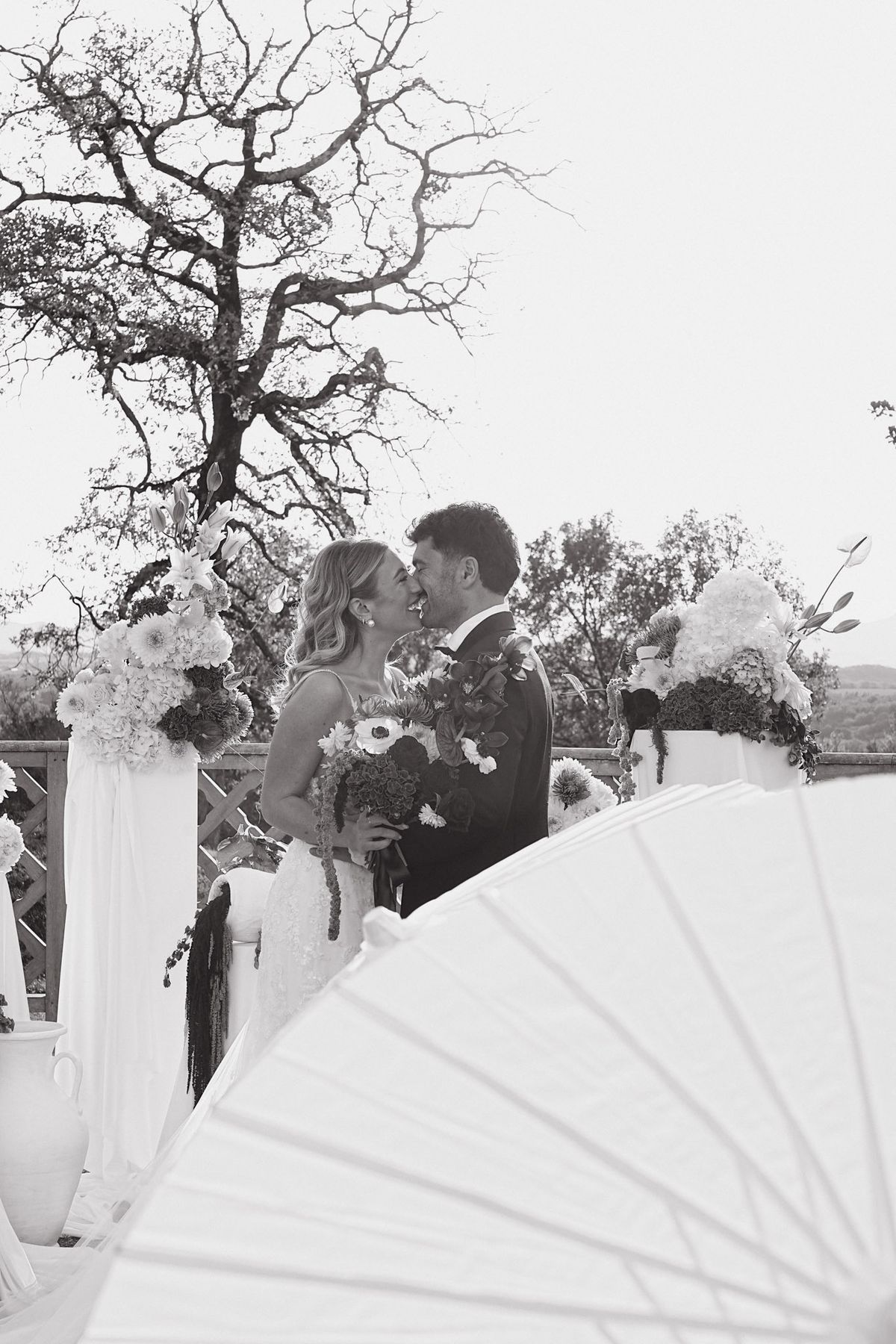 Bride and groom kiss at the outdoor ceremony arch, framed by floral pedestals, black and white