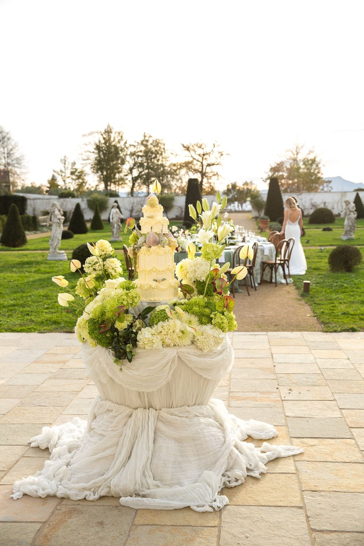 Garden cake plinth with crinkled white drape and overflowing florals leading to a reception table