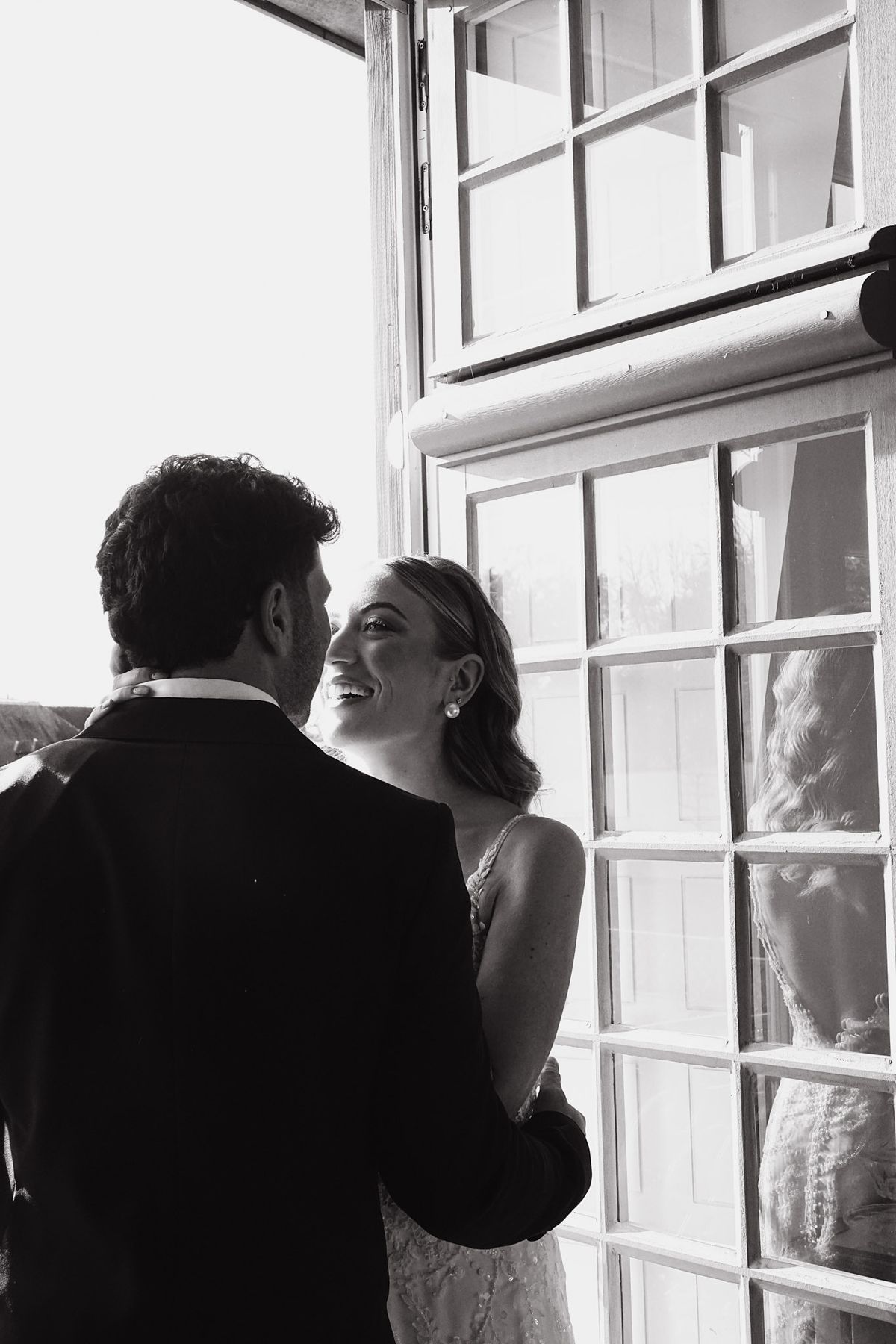 Couple share a quiet moment by a tall paned window, bride smiling up at the groom, black and white