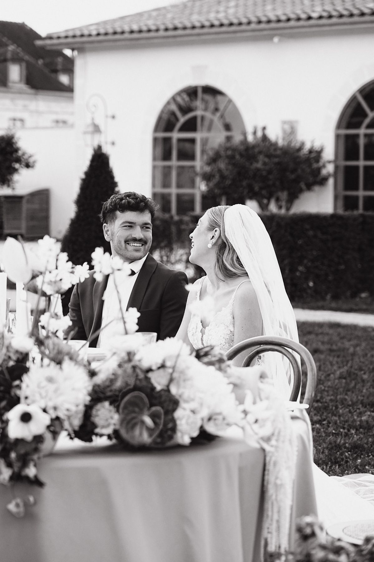 Couple laughing at the reception table with chateau arched windows behind them, black and white