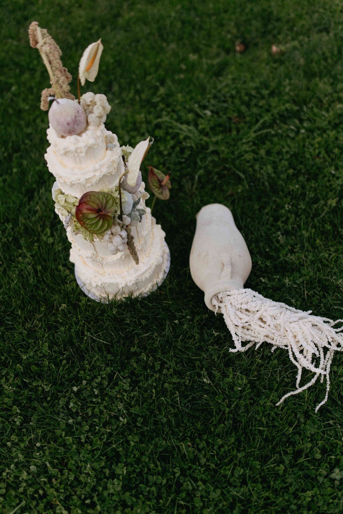Top-down view of a small tiered cake on grass beside a vessel spilling fringed cord