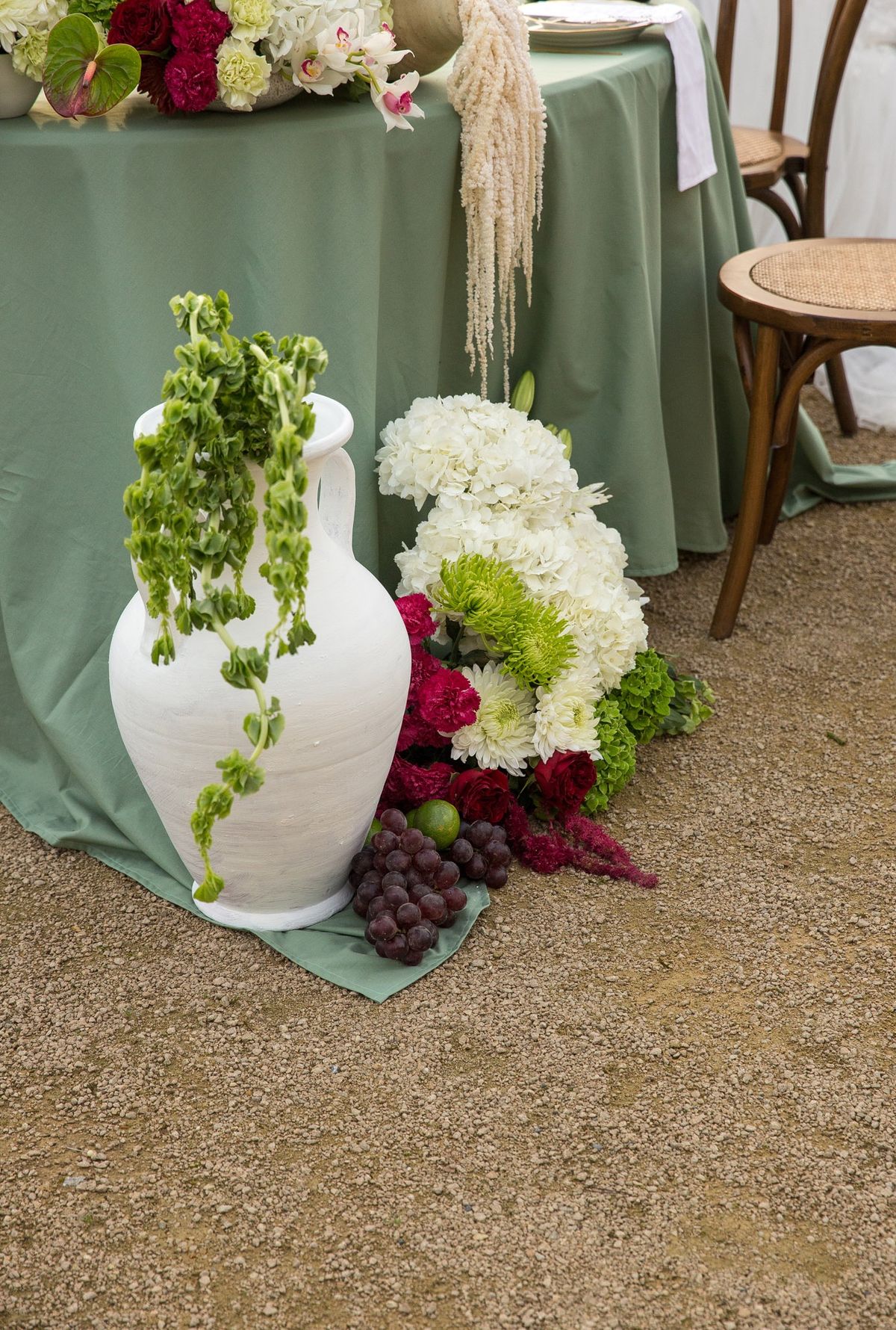 White amphora trailing greenery beside a sage draped table with hydrangeas and grapes