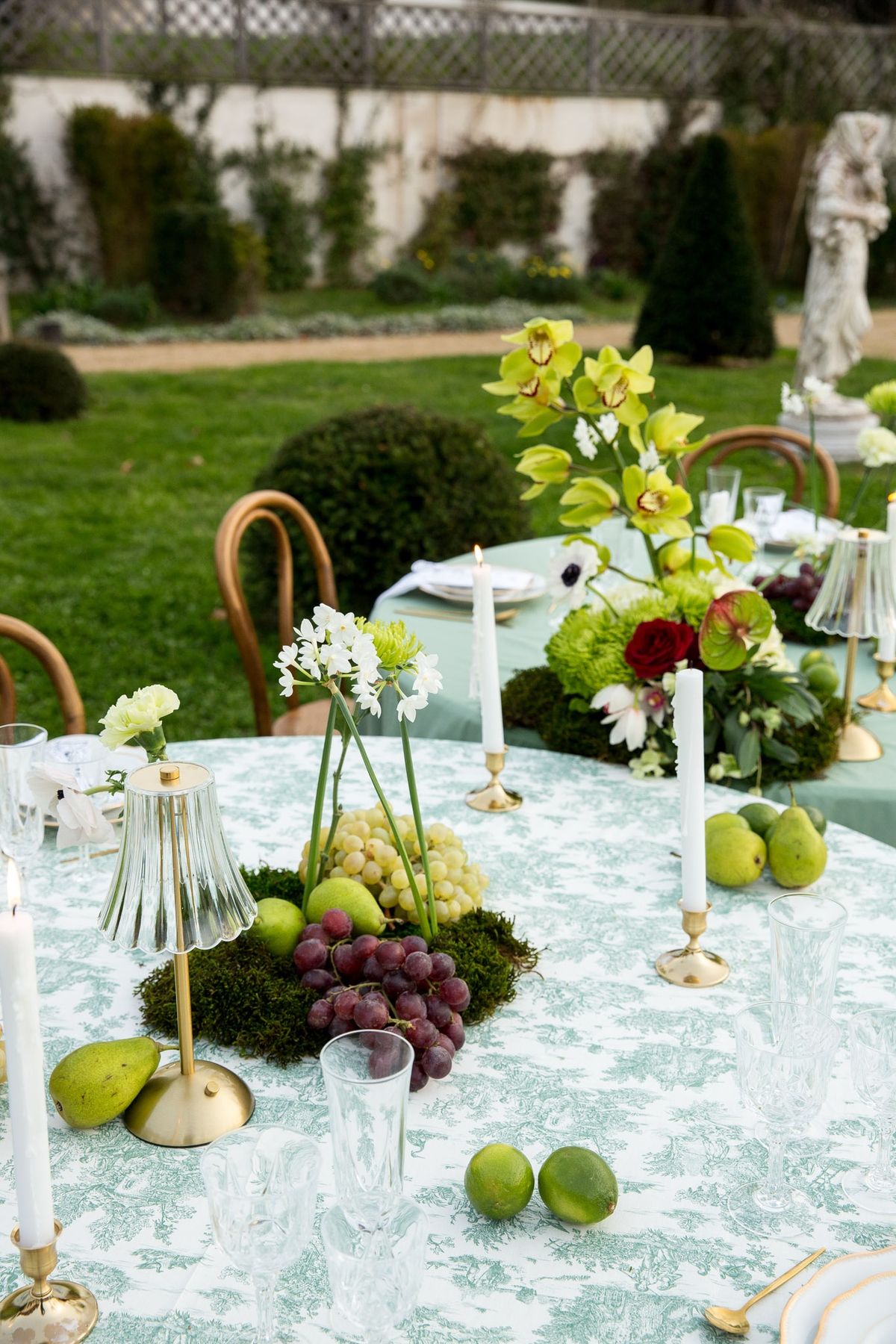 Round garden table with sage cloth, orchid centerpiece, fluted lamp and trailing grapes