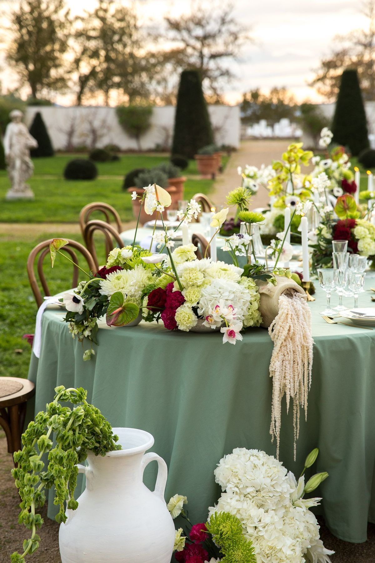 Round sage table with florals and amaranthus at twilight, garden statue and parterre behind