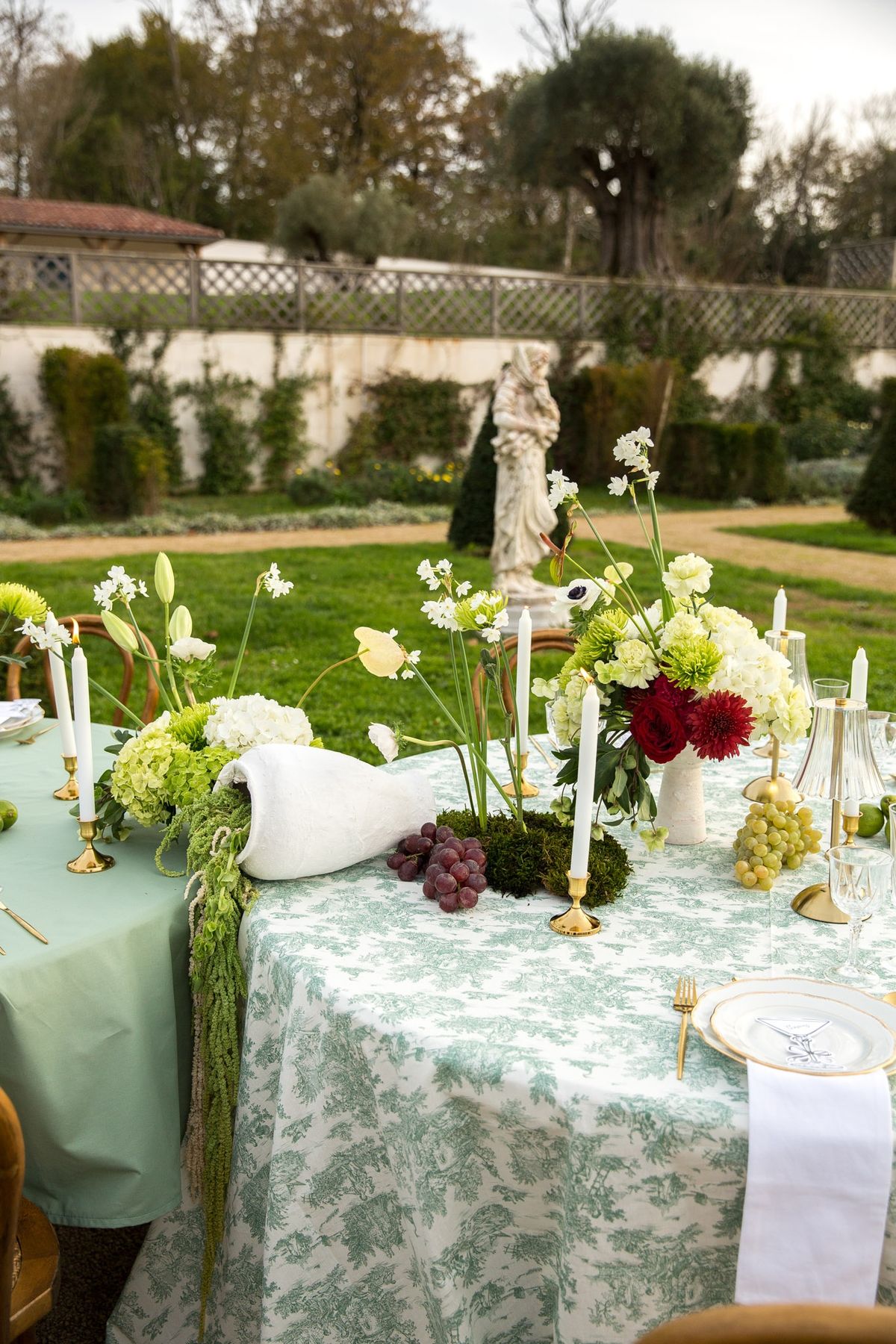 Wide twilight tablescape with round sage tables, statues and topiary along parterre path