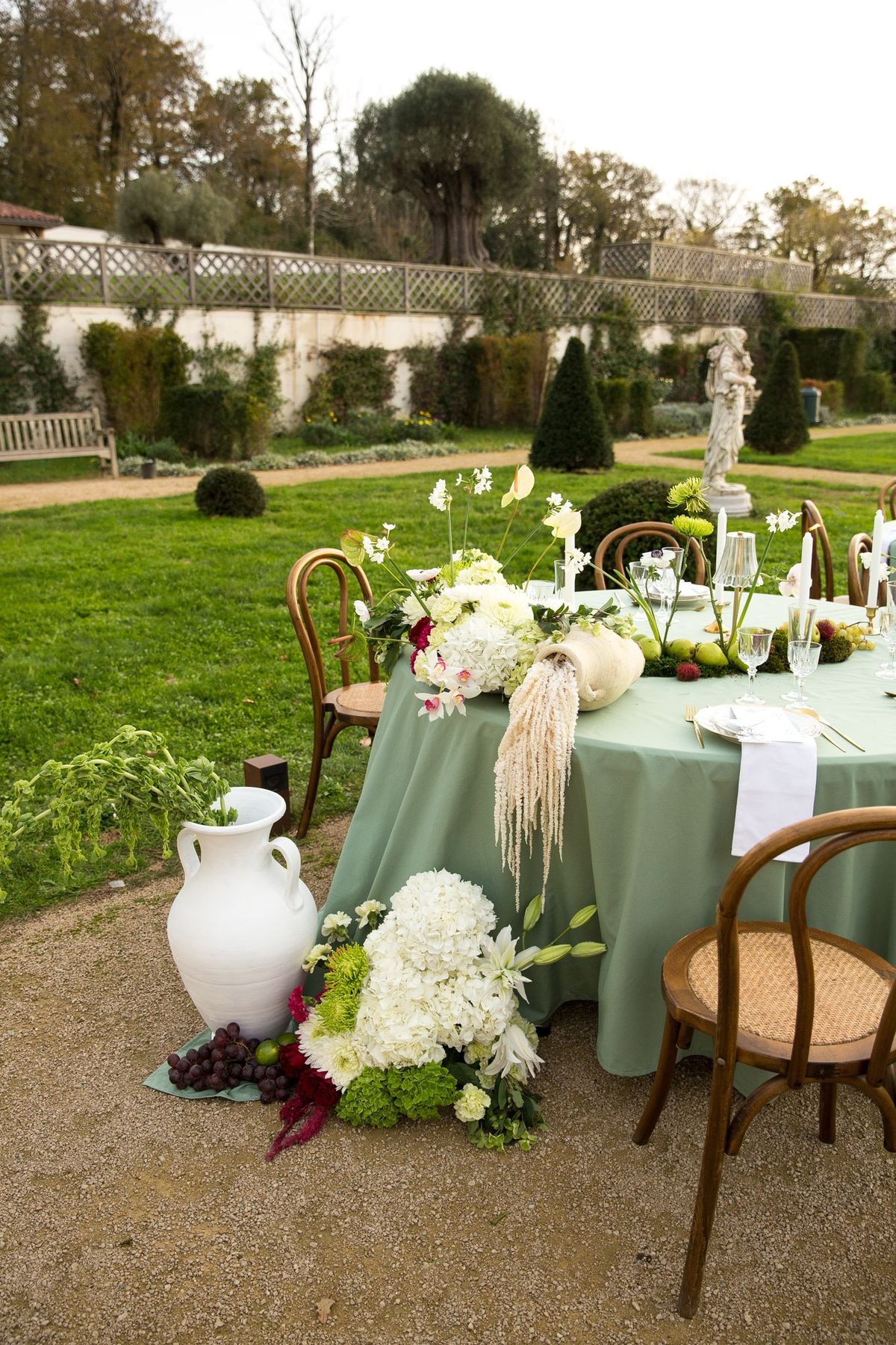 Sage round table with cascading floral runner, brass candleholders and garden statue behind