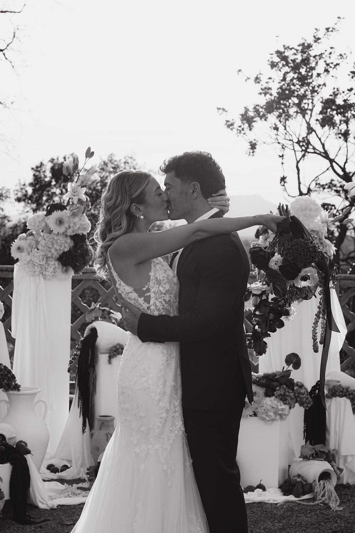 Couple kiss at the ceremony backdrop, bride raising her dark bouquet behind the groom's shoulder