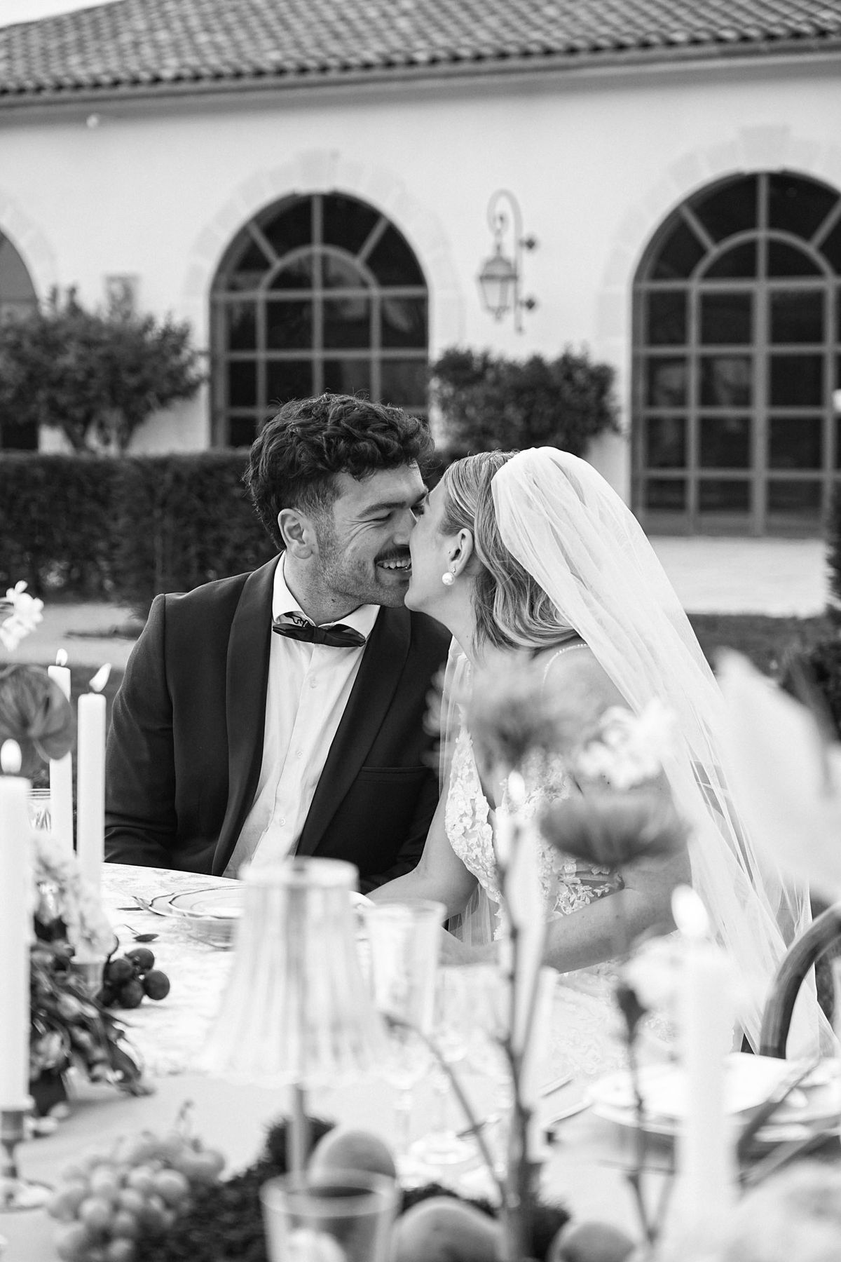 Couple touch noses at the reception table in black and white, chateau arched windows behind