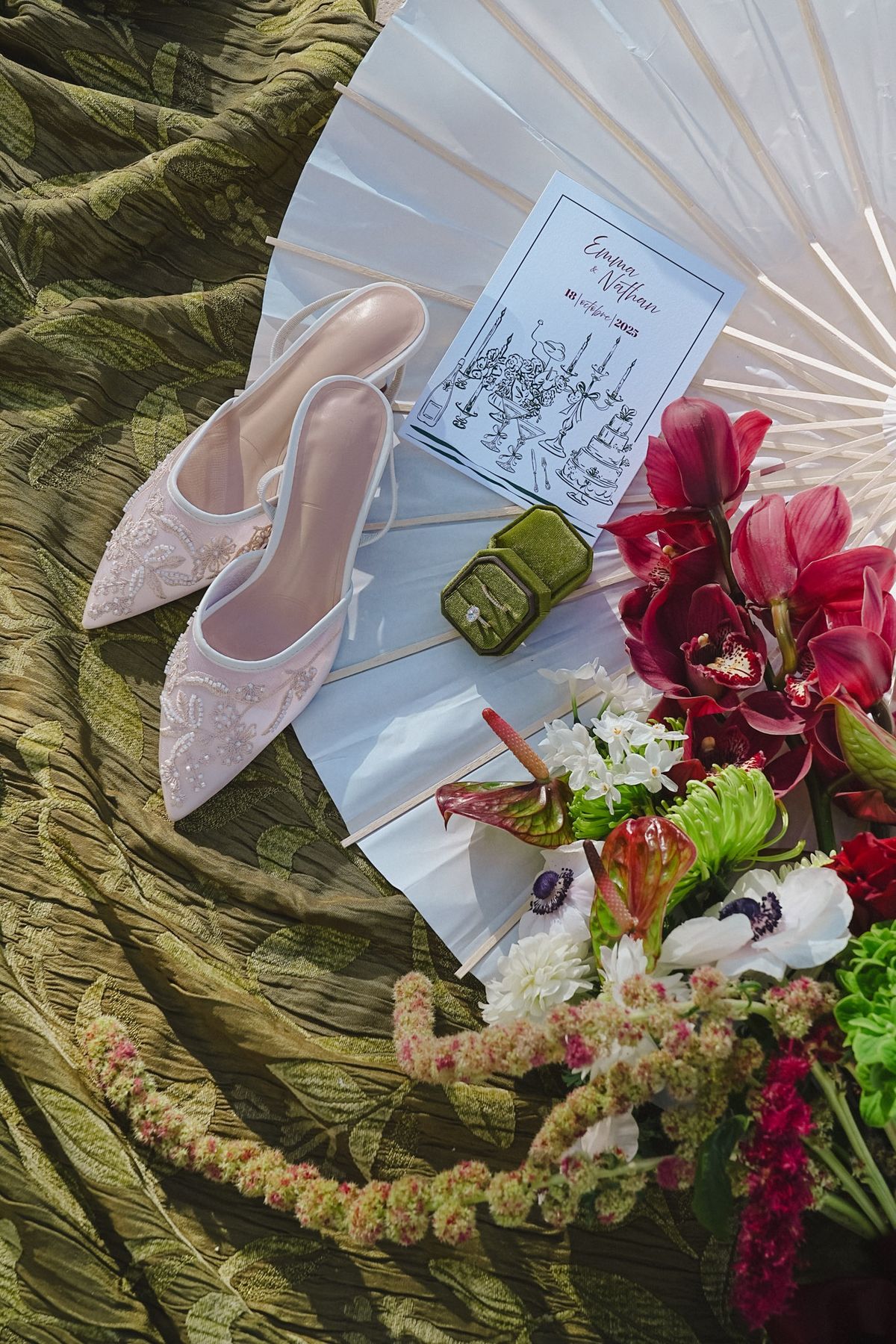 Flat lay with embroidered bridal mules, paper parasol, program, green ring box and floral posy