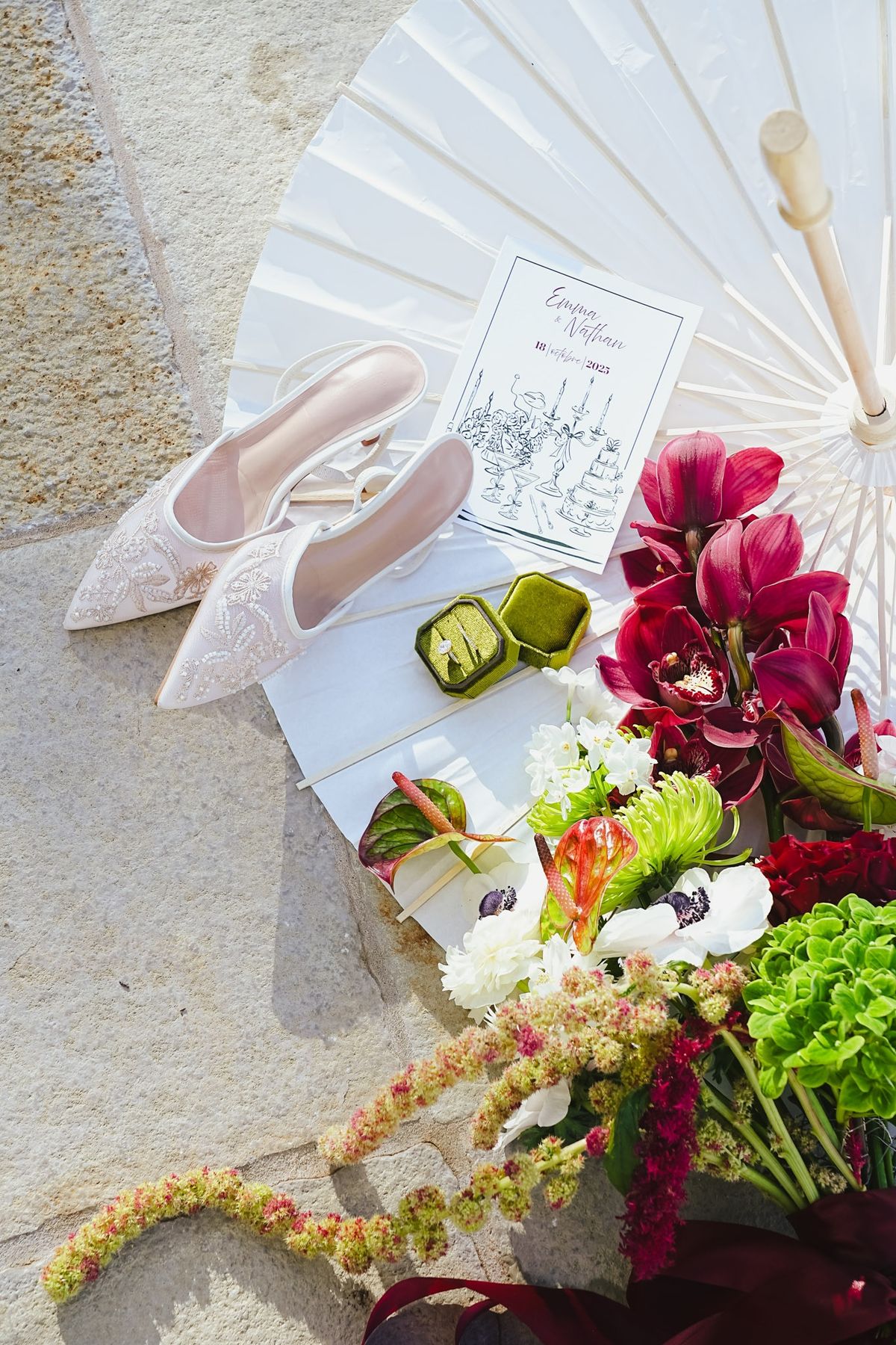 Flat lay on stone with bridal mules, paper parasol, program, ring box and posy of orchids and anemones