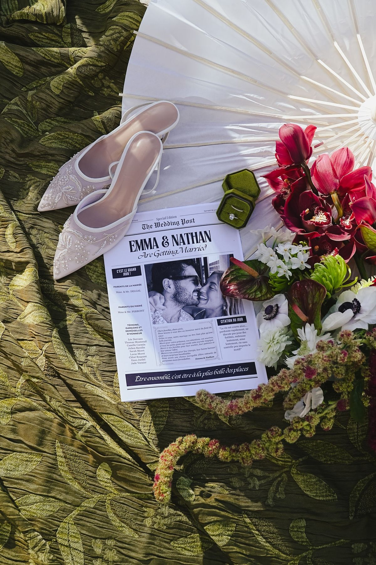 Newspaper style invitation flat lay with bridal mules, green ring box, posy and paper parasol
