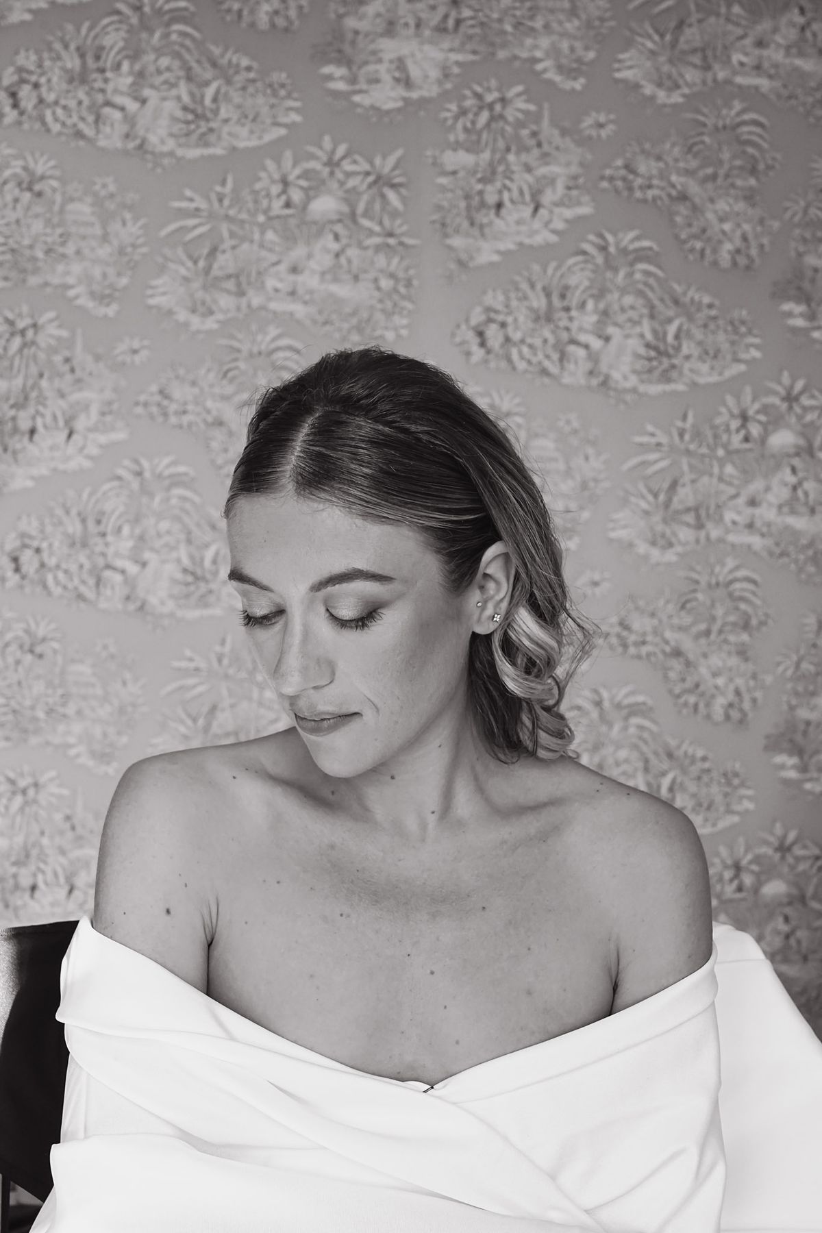 Bride looking downward in strapless wrap against printed toile wallpaper, black and white