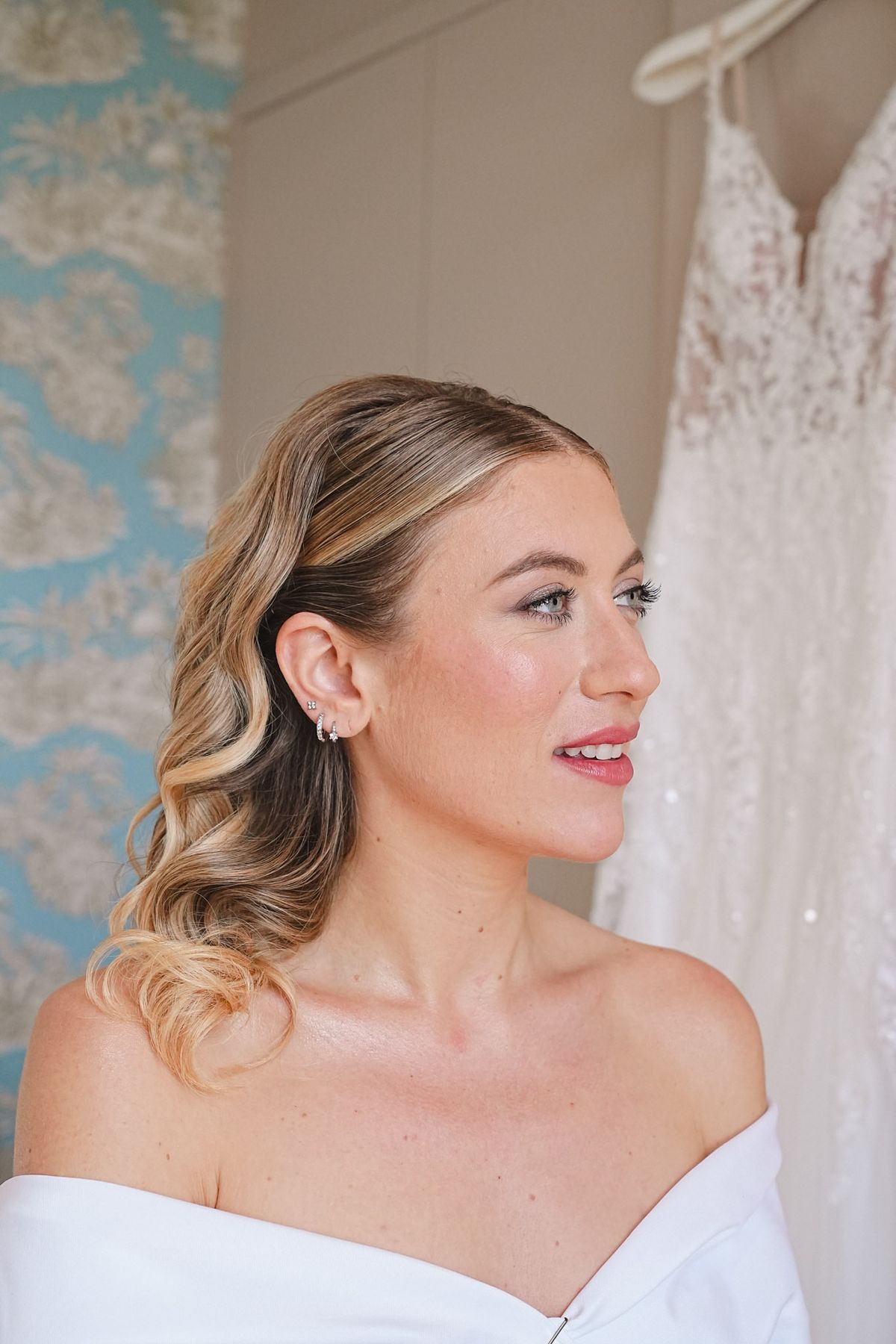 Bride in soft makeup with lace gown hanging behind her and turquoise toile wallpaper to her left