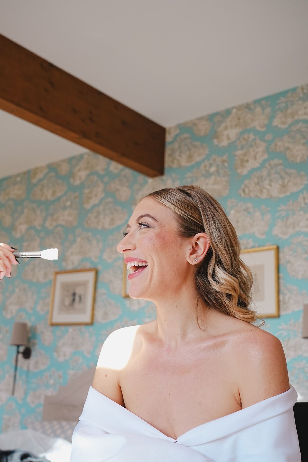 Bride laughing as a makeup brush enters frame, turquoise toile wallpaper and gilded frames behind