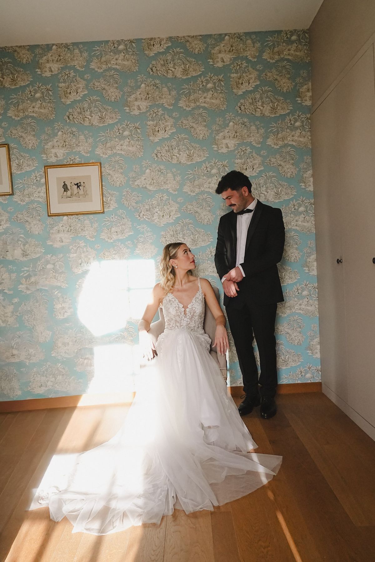 Couple in turquoise toile wallpapered bedroom, bride seated in tulle skirt with groom standing beside