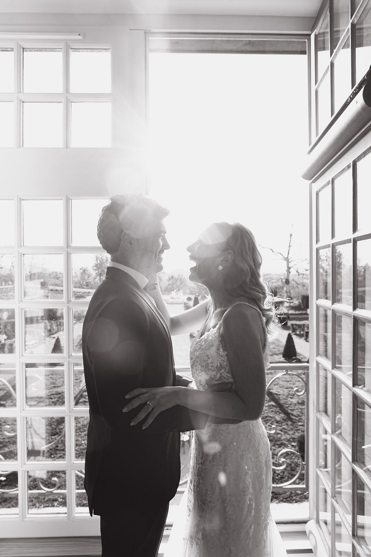 Couple silhouette in sunlit window, bride laughing as groom holds her waist, black and white