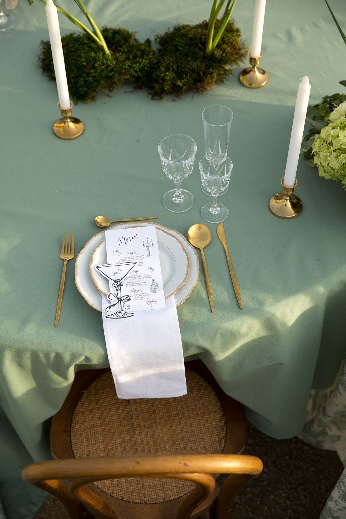 Top-down place setting on sage cloth with gold-rimmed plate, menu card, gold flatware and brass tapers