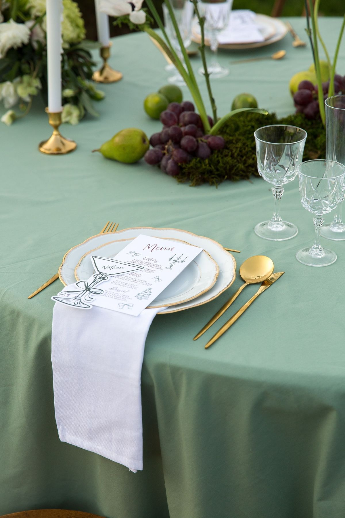 Close place setting with gold-rimmed plate, menu, white linen, gold flatware on sage cloth, fruit above