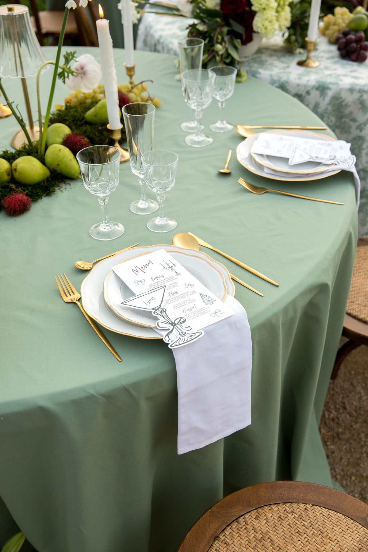 Tabletop with stacked plates, menu, white napkin and gold flatware on sage cloth, lamp and runner behind