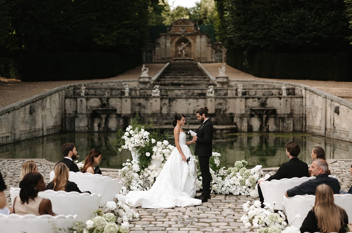 An outdoor wedding ceremony taking place on a cobblestone terrace in front of a grand ornamental stone fountain basin with classical statuary and a symmetrical staircase backdrop. The bride wears a strapless white ballgown with a full skirt and cathedral-length train, while the groom is dressed in a black tuxedo with bow tie and his hair in a bun; he is reading from a paper, likely his vows. The ceremony altar is flanked by large white urns overflowing with white peonies, ranunculus, and ferns, with additional ground-level arrangements of white blooms — including what appears to be baby's breath and hydrangea — lining the aisle. Guests, dressed predominantly in black formal attire, are seated in white ornate carved wooden chairs arranged on either side of the aisle, with approximately 20–30 visible guests. The overall styling is classic and monochromatic, with a strict white and green floral palette against the grey stone architecture. This is a wide editorial shot that frames the couple centrally with the full ceremony setting and architectural backdrop visible. Potential venue feature image.