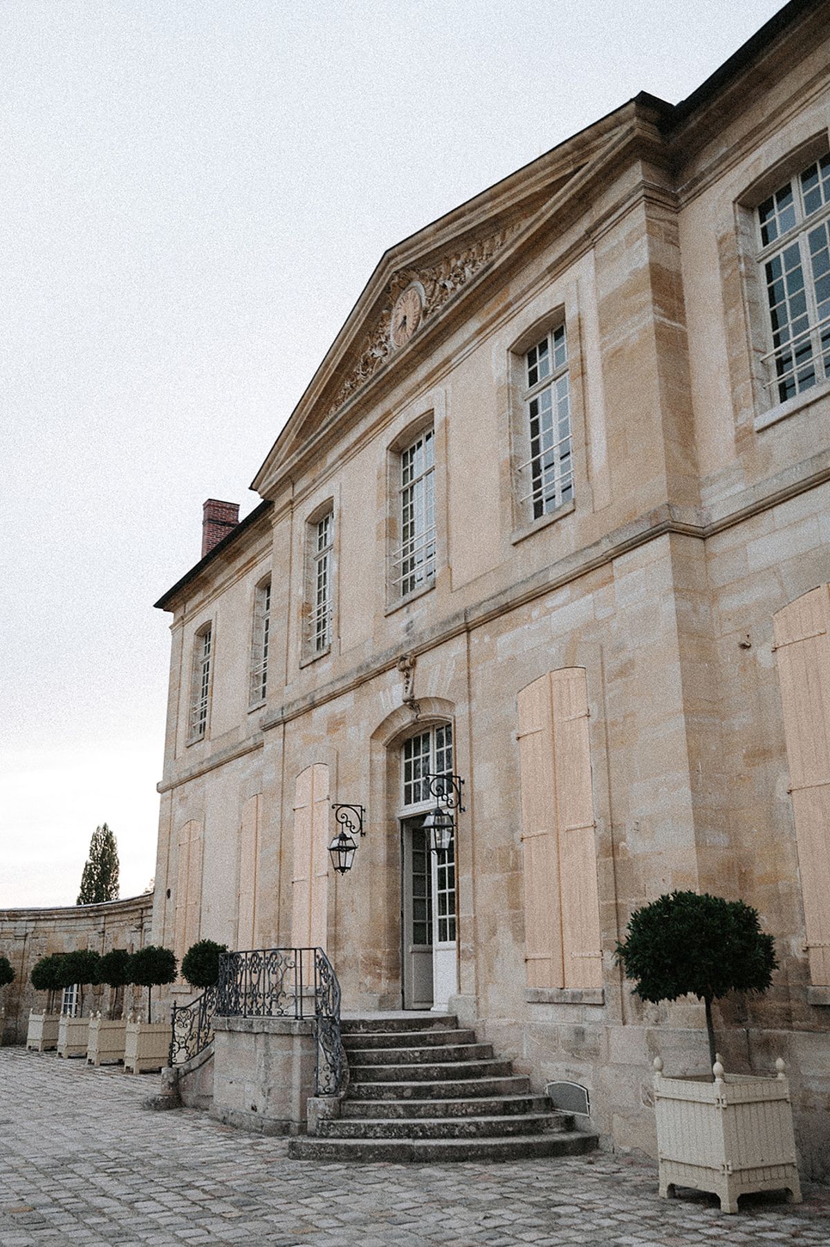 An exterior shot of a French château photographed from a low angle, showing the front façade in warm cream-toned limestone. The building features classical French architecture with a triangular pediment above the entrance bearing ornate carved detailing, tall multi-pane windows, and a central arched doorway flanked by two wrought-iron lanterns mounted on scrolled brackets. Stone steps lead up to the entrance door, bordered by a decorative wrought-iron railing. Cream-painted square planters holding neatly trimmed topiary balls line the forecourt on both sides of the entrance. The cobblestone courtyard extends in the foreground. The image has a soft, slightly faded film quality with muted tones. No people are present. Potential venue feature image.