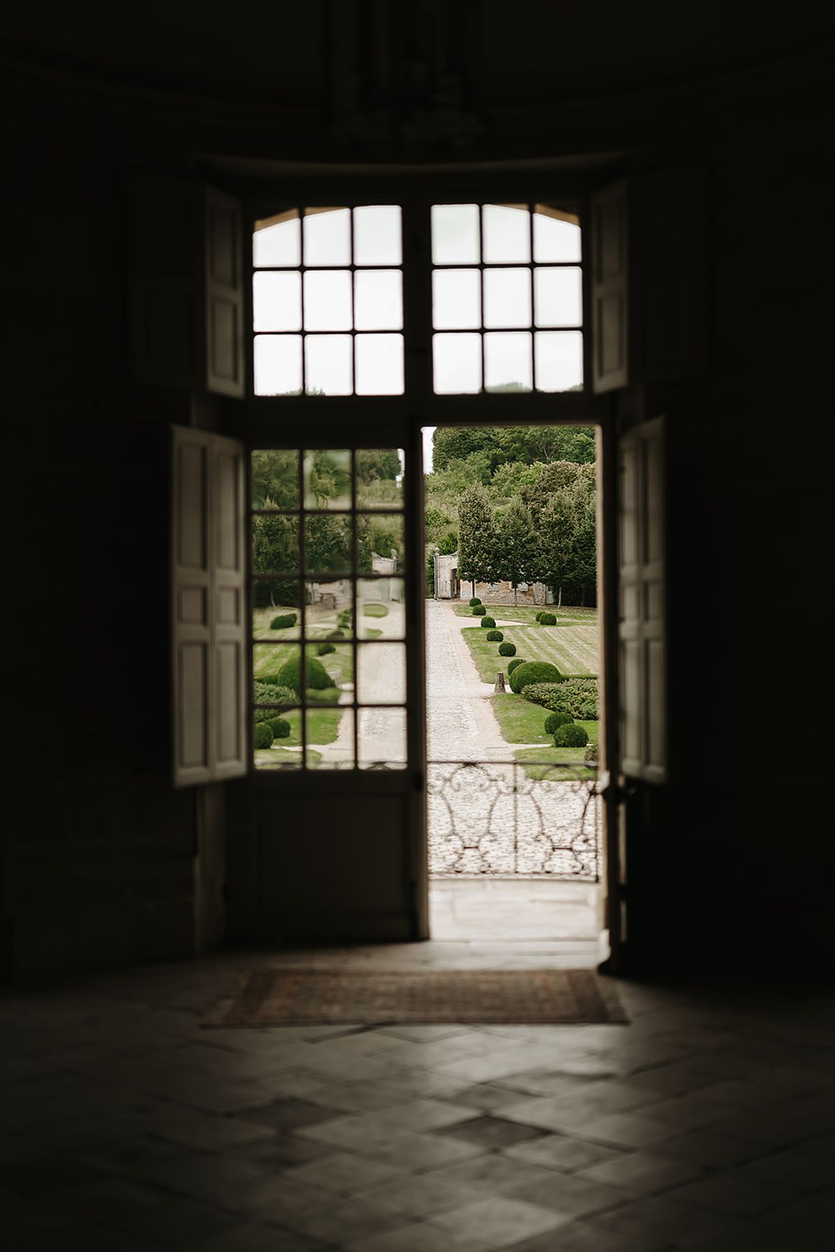 A venue detail shot taken from inside a château, looking outward through tall white-painted double doors with multi-pane glass panels. The doors are open, revealing a formal French garden with a gravel driveway, neatly trimmed spherical boxwood topiaries lining a central path, and a structured parterre garden to the left. An ornate wrought-iron railing is visible at the threshold, and the interior floor features stone diamond-pattern tiles. The foreground interior is in deep shadow, creating a strong contrast with the bright garden beyond. No people are present. Wide architectural shot framing the grounds as seen from within the building. Potential venue feature image.