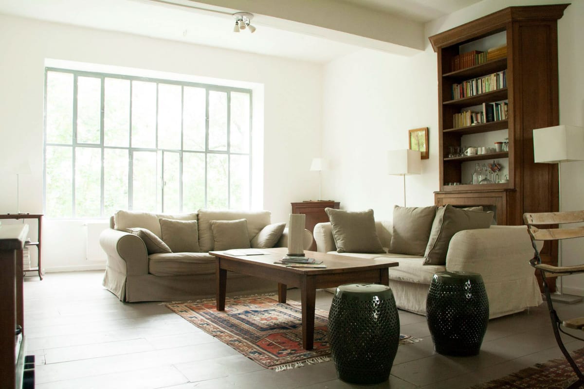 This image shows an interior living room or lounge space within what appears to be a venue property, with no wedding activity or people visible. The room features two linen-slipcovered sofas in a warm beige-grey tone arranged around a rectangular dark wood coffee table, atop a vintage red and blue patterned rug. Two dark green ceramic garden stool side tables are positioned in front of the seating area. A large multi-pane window with grey-green metal frames fills the back wall with natural light. A tall dark walnut bookcase filled with books and decorative items occupies the right side of the room, alongside a fireplace mantel. The decor palette is neutral — white walls, natural linen, warm wood tones — with a classic, understated country-house style. Wide shot. Potential venue feature image.