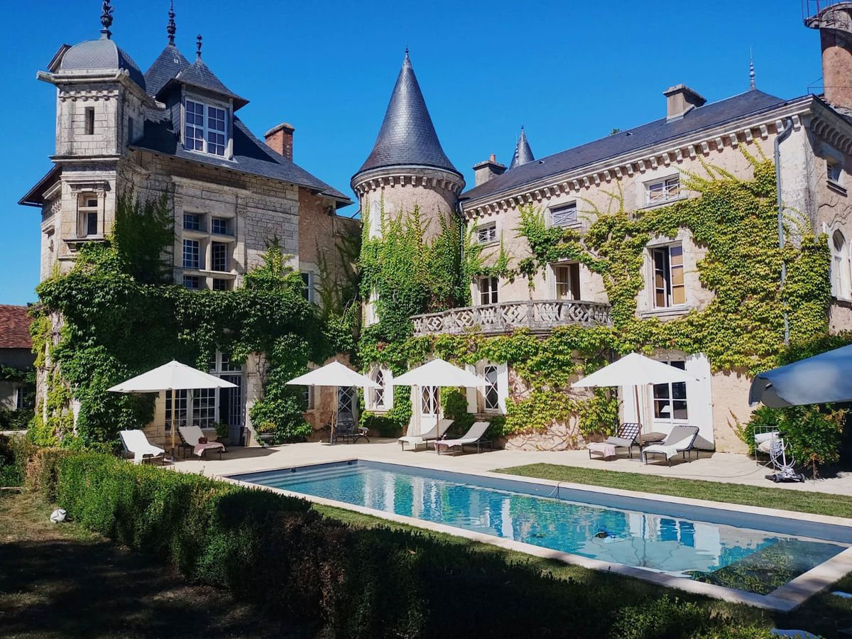 Wide exterior shot of a French château featuring multi-story limestone architecture with pointed slate-roofed turrets and a decorative wrought-iron balcony, extensively covered in climbing green ivy. In the foreground, a rectangular outdoor swimming pool is surrounded by a paved terrace furnished with white sun loungers and multiple white market umbrellas, creating a classic poolside setup. No people or wedding party are visible in the frame. Potential venue feature image.