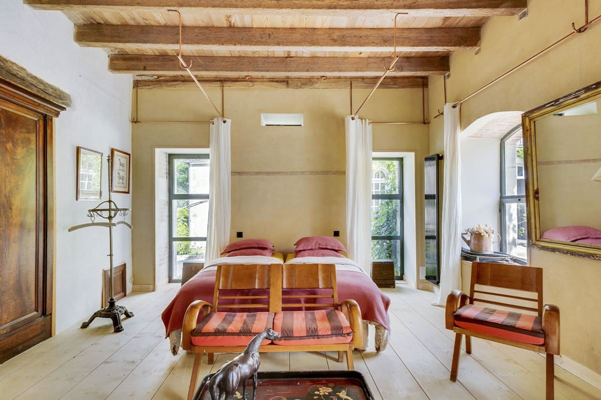 This is not a wedding photograph but rather an interior room shot, likely used as a venue accommodation or guest room feature image. The room is styled with a rustic-eclectic aesthetic, featuring exposed wooden ceiling beams, light-washed plank flooring, and warm ochre-toned walls. Two single beds with terracotta-red bedding and dusty rose pillows are positioned side by side, flanked by two large windows dressed with white linen curtains and copper curtain rods. The furnishings include mid-century wooden armchairs with orange and burgundy striped upholstery, an antique dark wood armoire, a brass floor lamp, and a large gilt-framed mirror. Decorative objects include a carved wooden elephant figurine on a lacquered tray in the foreground and a small dried floral arrangement on the windowsill. Wide shot taken from the foot of the beds. Potential venue feature image.