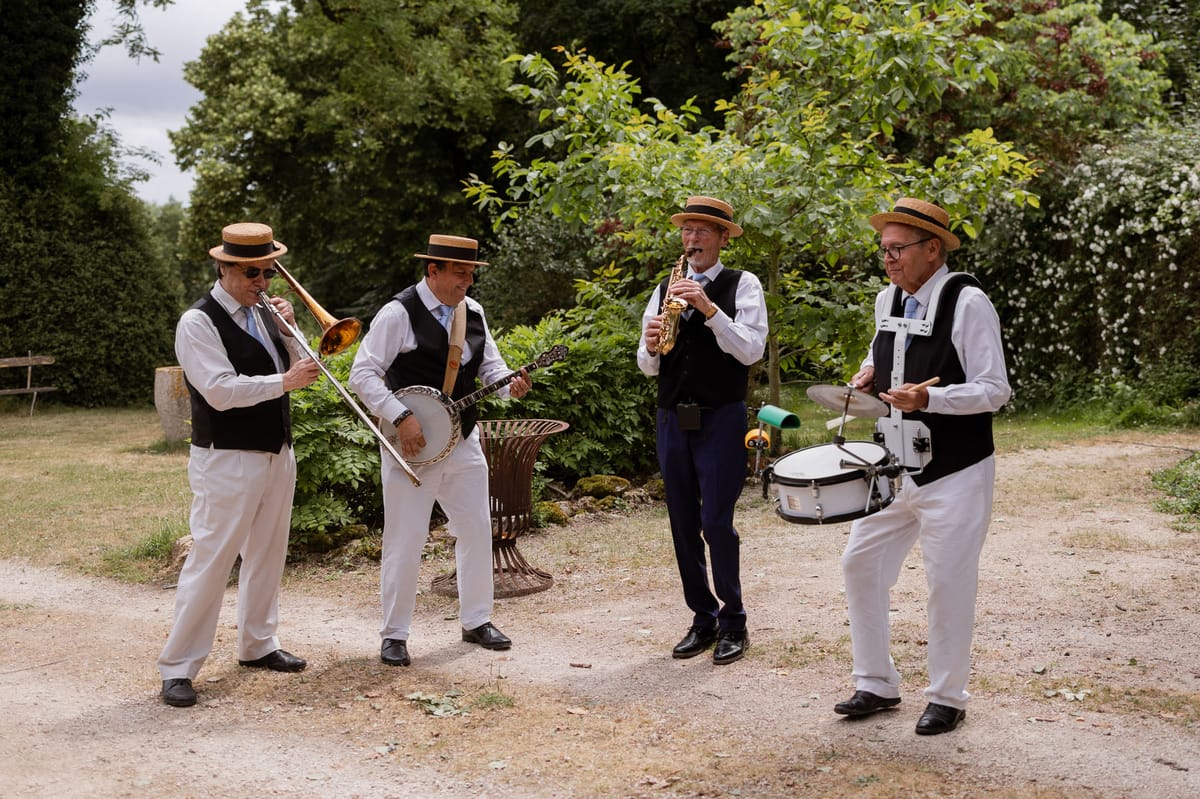 A four-piece live jazz band performs outdoors in a garden setting during what appears to be a wedding cocktail hour. The musicians are playing a trombone, banjo, alto saxophone, and snare drum, and are dressed in a coordinated vintage-style look: white trousers, black waistcoats over white shirts, light blue ties, and matching straw boater hats. Three of the four musicians wear sunglasses or glasses. The band stands on a gravel path surrounded by lush garden greenery. The wide shot captures all four performers in action, conveying an animated, upbeat atmosphere with a 1920s New Orleans jazz aesthetic.