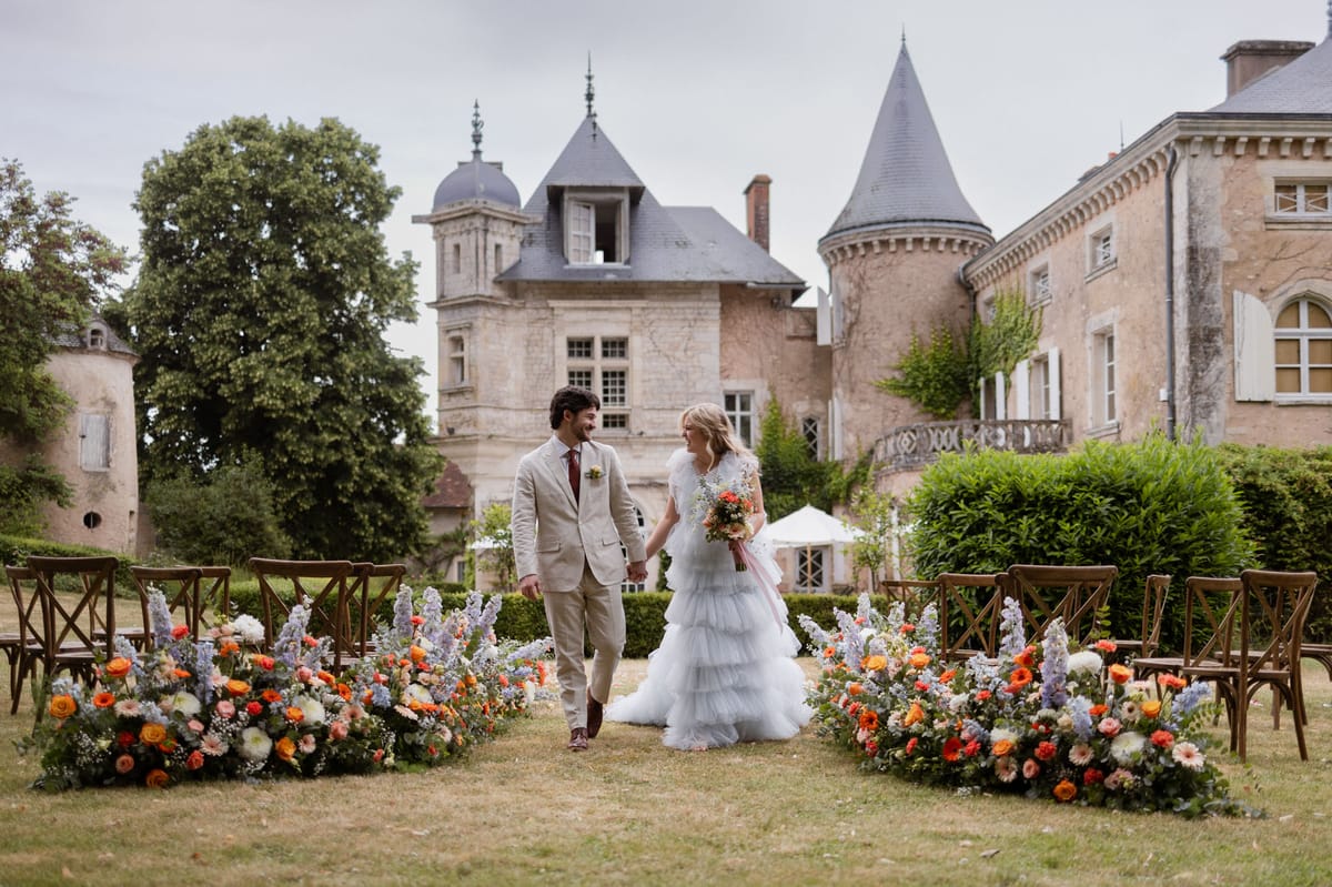 A couple walks hand-in-hand down an outdoor ceremony aisle set in the grounds of a French château, facing each other and smiling. The bride wears a multi-tiered ruffled tulle gown in pale grey-white with short sleeves, carrying a bouquet of orange ranunculus, blue delphinium, and mixed wildflowers with a blush ribbon; the groom wears a sand-coloured linen suit with a burgundy tie and a small floral buttonhole. The ceremony aisle is flanked by low floral ground arrangements featuring orange gerberas, orange roses, white dahlias, lavender-blue delphiniums, and eucalyptus foliage, with wooden cross-back chairs positioned on either side. The backdrop is a multi-turreted stone château with slate-grey conical roofs, set within formal hedged gardens. Wide portrait shot. Potential venue feature image.