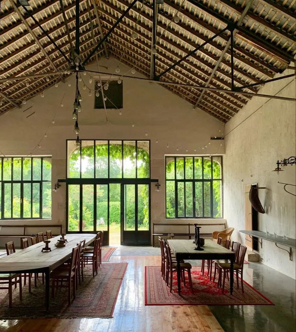 Interior shot of a converted barn or agricultural outbuilding set up for a wedding reception, with no people present. The space features a high vaulted ceiling with exposed wooden rafters and terracotta roof tiles, supported by dark metal beams. String lights with bare Edison-style bulbs are draped across the ceiling. Two long wooden farm tables with mismatched dark wood chairs are arranged on red patterned rugs over a mixed concrete and wood plank floor. A fireplace with a stone mantel and wall-mounted candle sconces are visible on the right wall. Large black-framed steel and glass windows with arched tops line the walls, and a central glazed double door opens onto a lush green garden. The overall styling is rustic-industrial with vintage furniture and warm, natural light flooding in from the garden-facing windows. Wide interior shot. Potential venue feature image.