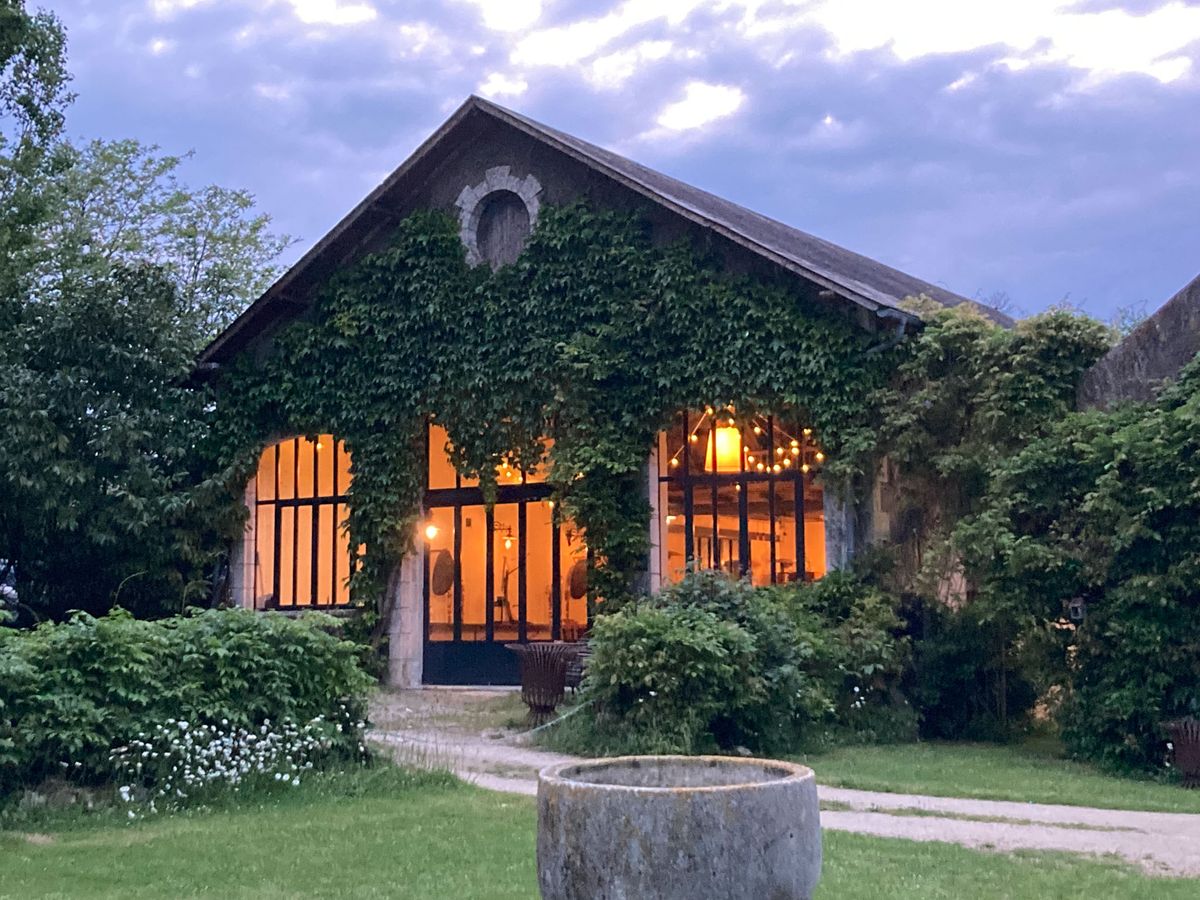 Wide exterior shot of a stone barn-style venue building photographed at dusk, with no people visible. The structure is heavily covered in green climbing ivy and features large black-framed industrial-style windows that glow with warm amber light from within, suggesting an event in progress inside. String lights are visible hanging inside the venue, visible through the windows and adding to the warm interior glow. The building has a pitched roof with a decorative oval stone window detail at the gable end. A gravel pathway leads to the entrance, flanked by a large weathered stone cylindrical planter in the foreground. Potential venue feature image.