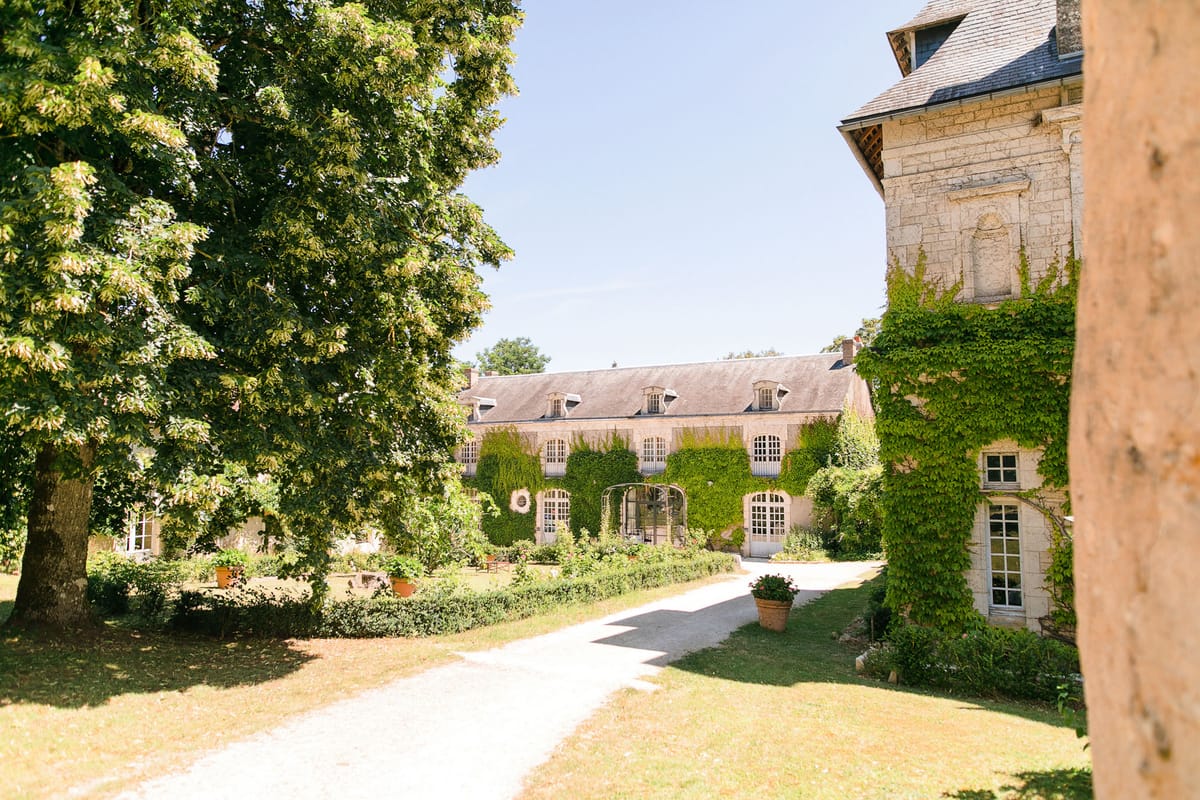 An outdoor wide shot of a French château estate taken in bright summer daylight. The image shows a long, two-storey limestone manor building with a slate mansard roof and multiple dormer windows, its facade heavily covered in climbing ivy and Virginia creeper. A gravel courtyard path leads toward the building's central arched glass conservatory entrance, flanked by terracotta pots with plantings. A second ivy-covered stone tower or wing is visible in the foreground right. No people are present in the frame. Potential venue feature image.